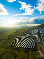 Solar panels cover a large area in a valley under a clear blue sky as the sun sets in the background near a mountain