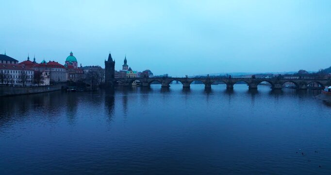Cold blue winter morning in Prague with a distant view of Charles Bridge over the Vltava River. Tourists cross the historic landmark in a calm, frosty atmosphere.