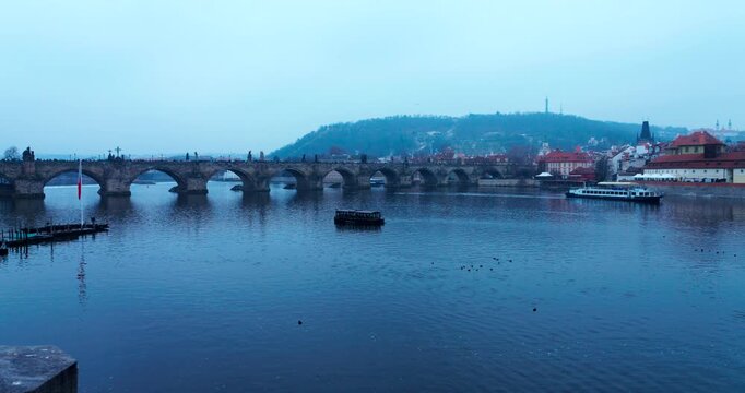 Cold blue winter morning in Prague with a distant view of Charles Bridge over the Vltava River. Tourists cross the historic landmark in a calm, frosty atmosphere.