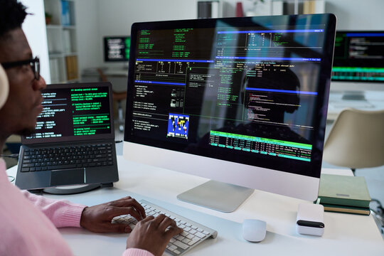 Black young man working on desktop computer with multiple programming code windows open on large monitor, using keyboard and mouse, laptop with code in background