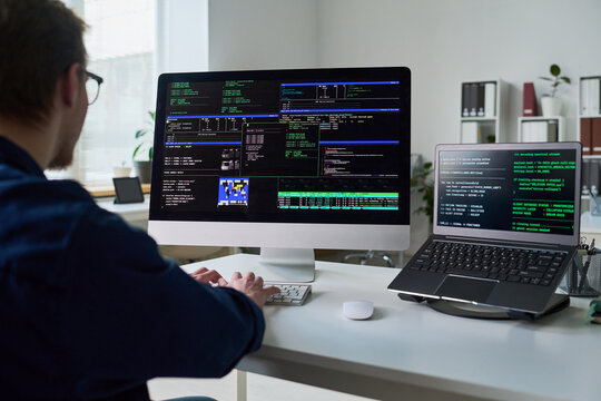 Young man working at desk using desktop computer and laptop displaying programming code, sitting in office environment, focusing on software development tasks