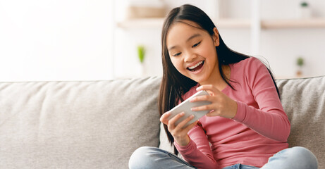 A girl sits on a couch in a bright room. She is smiling while looking at her smartphone, showing...