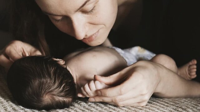 A woman leans over a newborn baby, gently stroking and kissing its tiny hands while lying on a textured blanket indoors