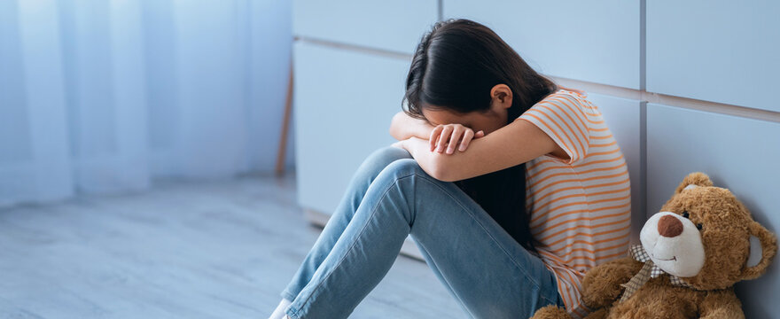 A young girl sits on the floor in a room with wooden flooring. She has her arms covering her face. A teddy bear is nearby. Sunlight enters through a window and illuminates the space.