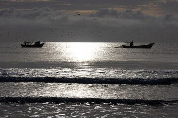 Landscape composed of two boats at sea at sunrise; sunlight reflects on the water, creating a silhouette effect on various elements