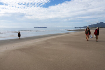 Two women walking on a beach on the southern coast of S&atilde;o Paulo and another woman heading towards the water; the sky is clear with clouds
