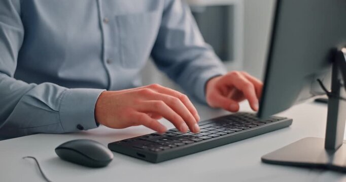 Close-up of adult male hands and forearms typing on black slim keyboard wearing light blue dress shirt with buttoned cuffs, white desk with dark wireless mouse in professional office