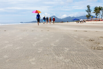 The image shows a group of people walking on a beach in the center of the image, with mountains in the background and other people engaged in various activities on the beach