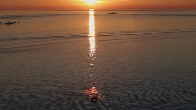 Clean horizon sunset shot with glowing sun trail across smooth ocean surface near Lisbon. Catamaran facing the sun and the Bugio lighthouse