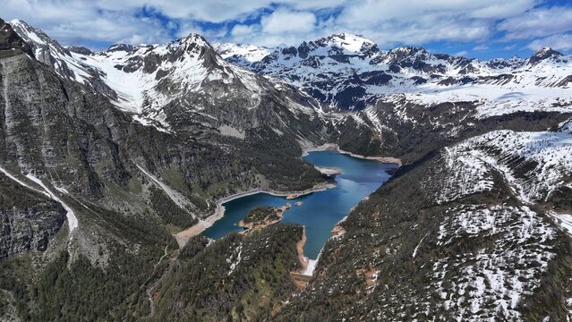 Aerial view of a sapphire lake nestled amidst rugged, snow-capped mountains, a stark contrast to the verdant valleys below, Alpe Devero, Piemonte, Italy.