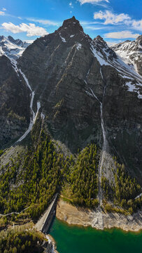 Aerial view of a dramatic mountain peak, its rugged slopes cascading down to a turquoise lake, contrasting with patches of snow and lush greenery, Alpe Devero, Piemonte, Italy.