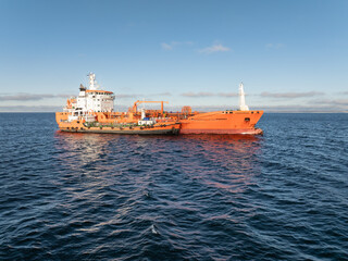 Fototapeta premium Aerial drone view of a bright orange industrial chemical tanker ship performing bunkering operations in the blue waters of Baltic Sea.