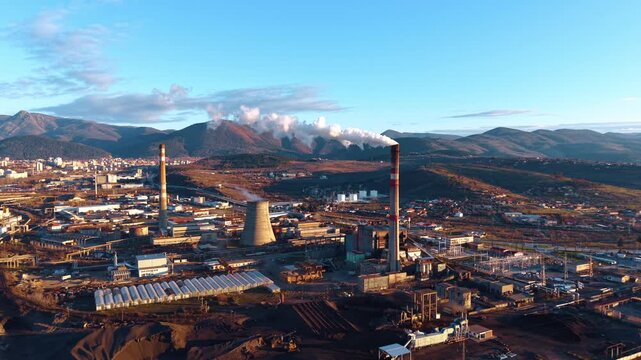 Wide panoramic view of industrial zone and power station. Extensive industrial area with smoking chimneys, cooling towers and power lines at sunset near mountains.