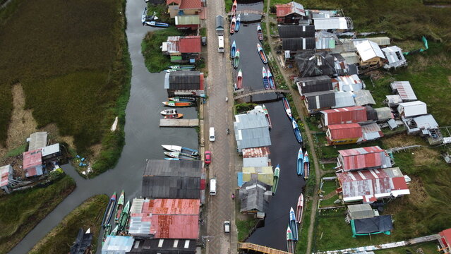 Drone footage flying over a small village at Laguna de la Cocha, Colombia. Traditional boats lined along the waterway and houses beside the lagoon in a rural landscape.