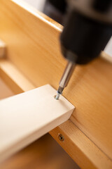 Worker uses drill to secure screw into wooden furniture part during assembly process in a home workshop