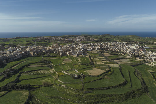 Aerial view of sun-drenched fields meeting the ancient walls of Gharb under a vast sky, a tapestry of green and stone unfolds before the azure sea, Gharb, Ta Pinu bazilika, Malta.