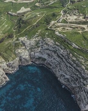 Aerial view of the rugged coastline where turquoise waters meet towering cliffs, creating a striking contrast against the lush green landscape, Dwejra Bay, San Lawrenz, Malta.