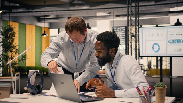 Diverse corporate office scene with team collaboration around laptop and reports, research and development. Professional people analyzing KPI metrics and insights for business intel.