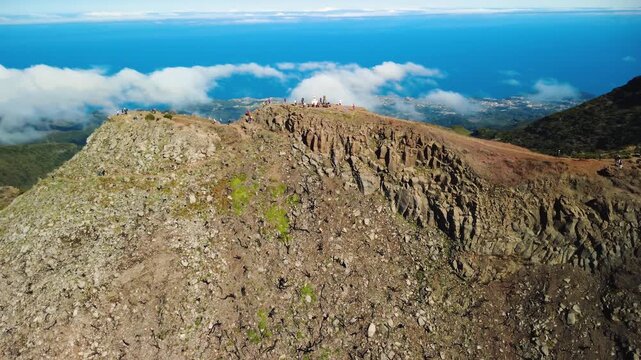 Aerial orbit of Pico Ruivo peak, drone rotating to the south revealing deep volcanic valleys and steep ridges, hikers at the summit marker under cinematic sunlight in Madeira highlands, 4K.