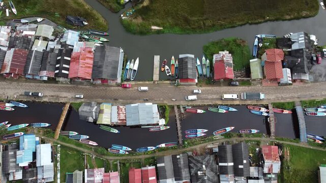 Drone footage flying over a small village at Laguna de la Cocha, Colombia. Traditional boats lined along the waterway and houses beside the lagoon in a rural landscape.
