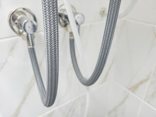 Braided stainless steel water supply hoses connected to bathroom valves on white tile wall, close-up.