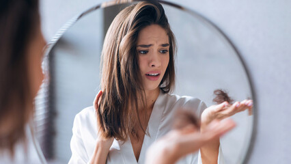 A woman stands in front of a mirror looking surprised while holding a clump of hair in her hand. She seems concerned as she examines the hair loss during her morning routine. © Prostock-studio