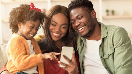 Fototapeta premium A young family sits together on a couch. The child points at a phone while the parents smile and look closely at the screen. They share a moment of joy in a warm home setting.