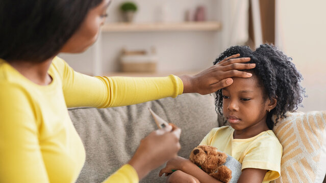 A mother checks the temperature of her sick child who is holding a teddy bear while sitting on a couch. The room is bright with soft furnishings around. It is a comforting moment.