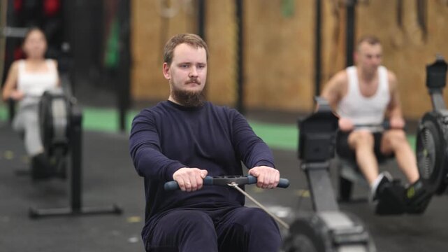 Motivated sporty young adult bearded man doing intense strength training, working out on rowing machine in modern gym. High quality 4k footage