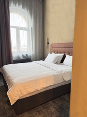 One large double brown bed with white bedding and pillows in a room with a window behind a curtain in the bedroom