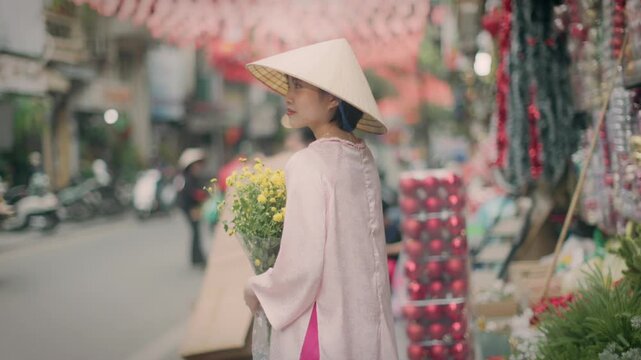 In Hanoi, Vietnam, a woman in traditional dress and a conical hat holds yellow flowers on a busy street. The image captures the essence of Vietnamese culture.