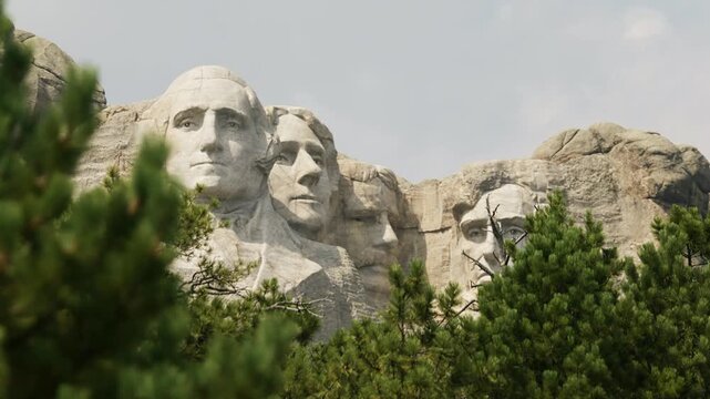 Steady shot of Mount Rushmore National Memorial, featuring pine trees in the foreground; bright sky.