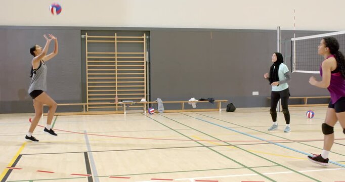 Diverse women's team tossing volleyball in school gym setter leaping at net blocker blocking rally