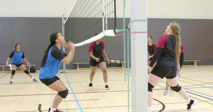 After female setter sending pass over net, blue-jerseyed woman blocking ball with knee pads at gym