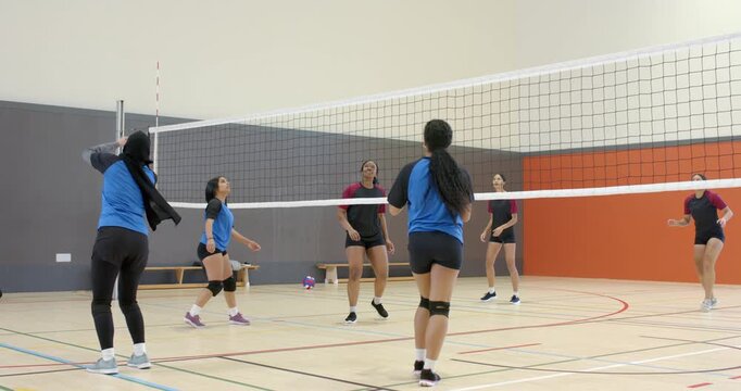 Hijab-clad setter reacting to descending ball, lobbing it over net while rivals bumping back at gym