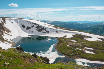 Scenic sunlit landscape with alpine lake in rocky snowy cirque near stone hill top during thaw. Ice floats in mountain lake among rocks and snows with view to forest mountain range under cloudy sky. © Daniil