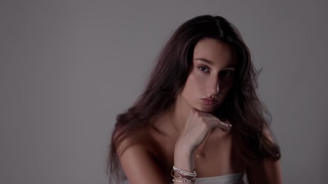 Studio portrait of a young woman with long brown hair, wearing bracelets and a necklace, posing with her hand on her chin against a gray background
