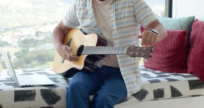 Man is turning tuning pegs and plucking acoustic guitar on window bench while following laptop