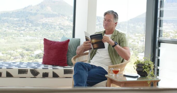 Mature man sitting armchair reading booklet shifting grip glancing at hillside view, copy space
