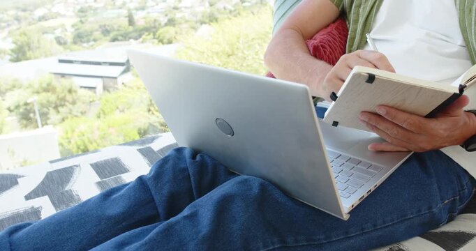 Person angling laptop forward and flipping notebook, then writing notes for work on patio blanket