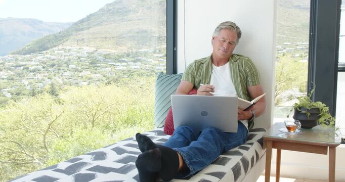 Man reclining on daybed at home with laptop open, noting notebook with pen, working remotely