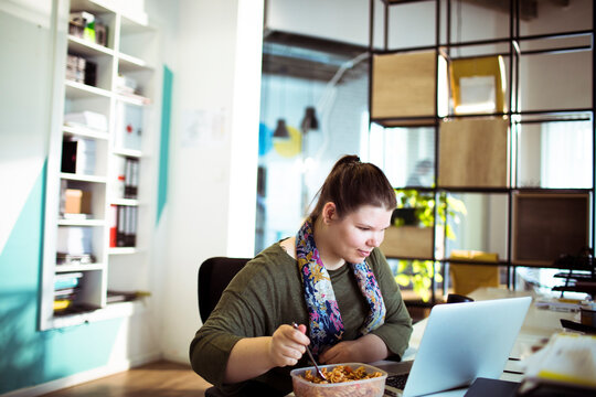 Young woman eating lunch while working on laptop in modern office