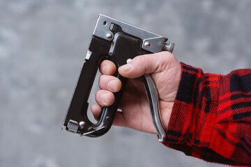 Close up of male hand in red checkered work shirt holding black manual staple gun against neutral gray background. Concepts of construction and DIY home renovation.