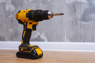 Professional cordless electric drill screwdriver standing on laminate floor against grey concrete wall background.