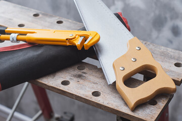 Yellow adjustable pipe wrench and classic crosscut hand saw with wooden handle rest on a wooden workbench. Concept professional handyman toolkit. Construction tools and workshop setups.