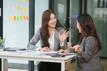 Fototapeta premium Two Businesswomen office colleagues analyzing data on a laptop and tablet, sharing ideas with smiles, and celebrating their success together in a modern office.