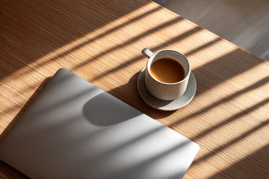 Closed aluminum laptop and coffee cup on oak desk with sunlight shadows