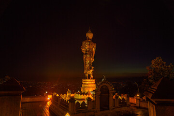 Fototapeta premium A golden standing Buddha faces the rising sun at Wat Khao Noi, glowing gently as morning light spreads across the horizon above Nan city.