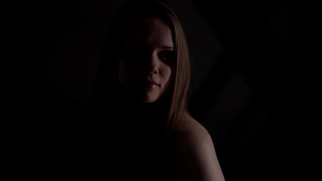 Portrait of a young Caucasian woman with long brown hair looking down, illuminated by a soft light in a dark room