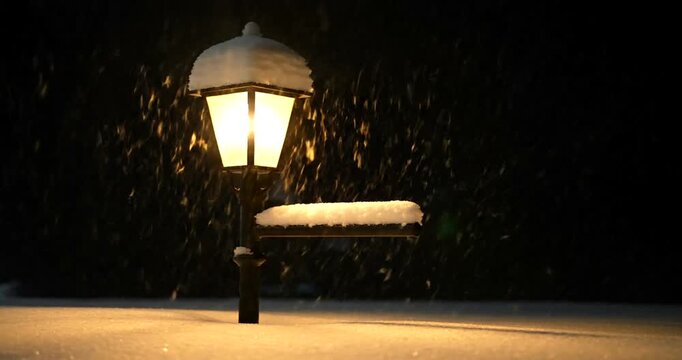 Snowy street lamp and bench illuminated in a serene winter snowfall scene at night, viewed from the front.